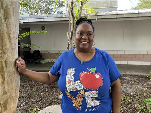 Ms. Rodriguez standing in courtyard next to a tree wearing a blue t shirt with a red apple on it